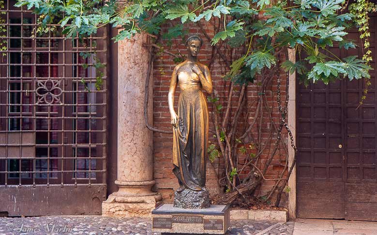 A gold coloured female statue at Casa Di Giulietta, Italy, in front of an old building with a tree branch above her