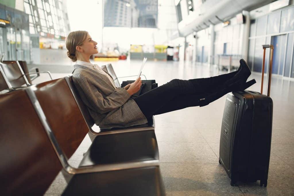 A lady sitting and waiting at the airport with her legs on her luggage while holding a book.