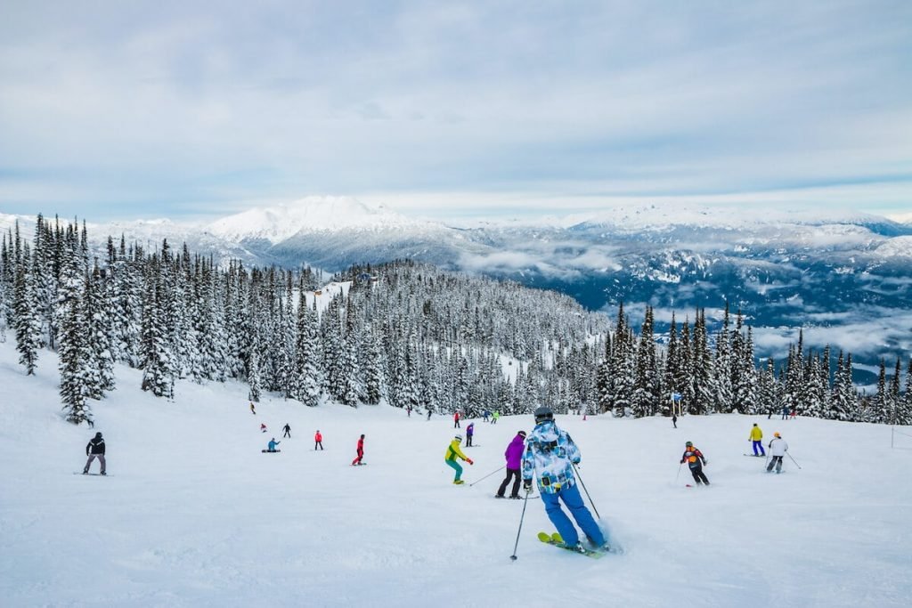 A group of people skiing in a large snow covered area with mountains and snow covered trees around them