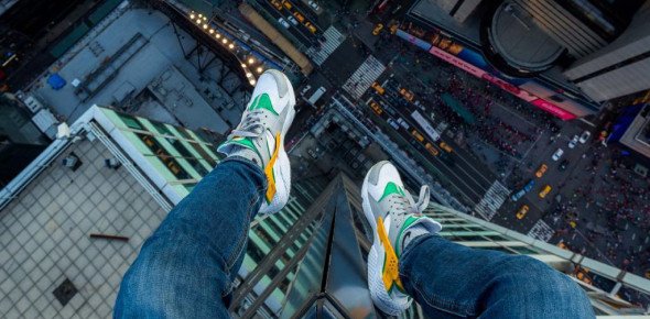 A male's foot with sneakers and a pair of jean sitting on a very tall building, overlooking a busy city.