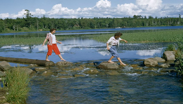 Two Ladies walking over a fresh water lake by stepping on some piles of stones and woods