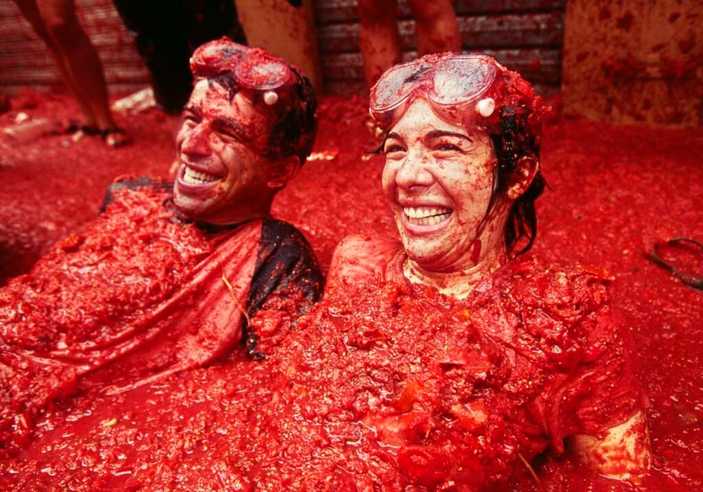 A lady and a man smiling while being drenched with red tomatoes